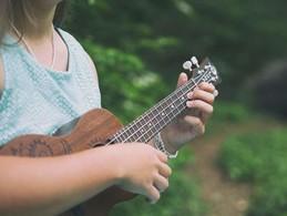 picture of a woman in a blue shirt against an outside green background strumming a ukulele